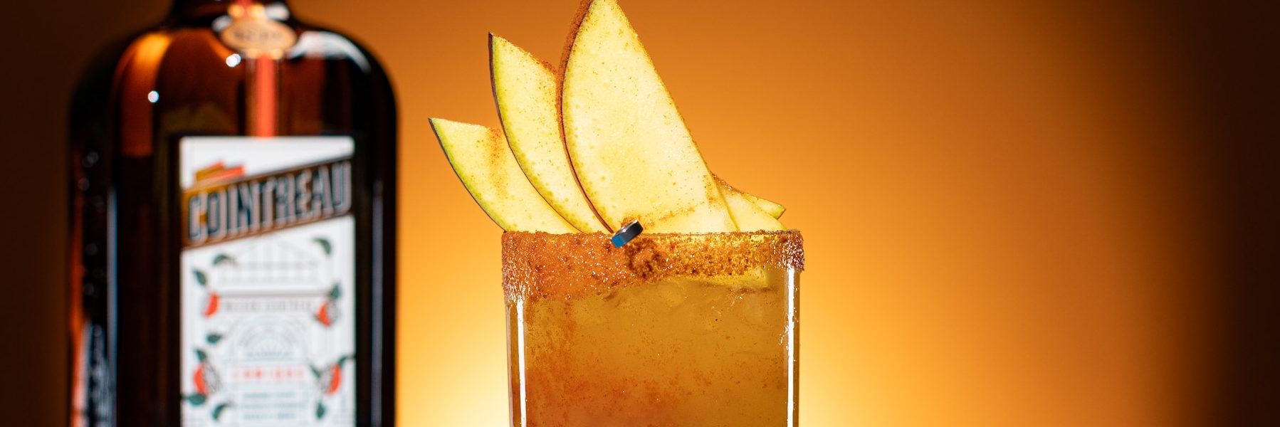 Cointreau Spicy Mango Margarita in short glass with apple garnish and Cointreau bottle on orange background