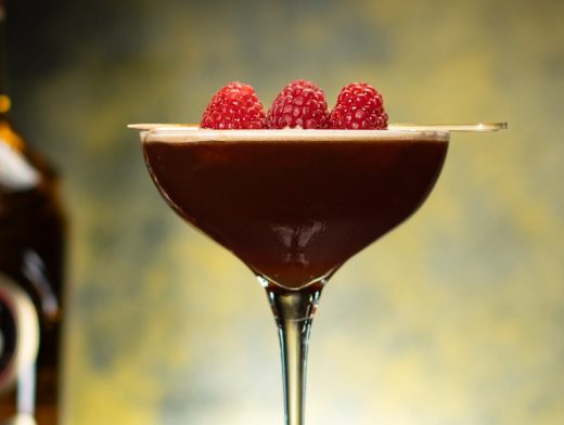 Licor 43 Deja Brew 43 in a coupe glass garnished with raspberries with licor 43 on yellow and black background