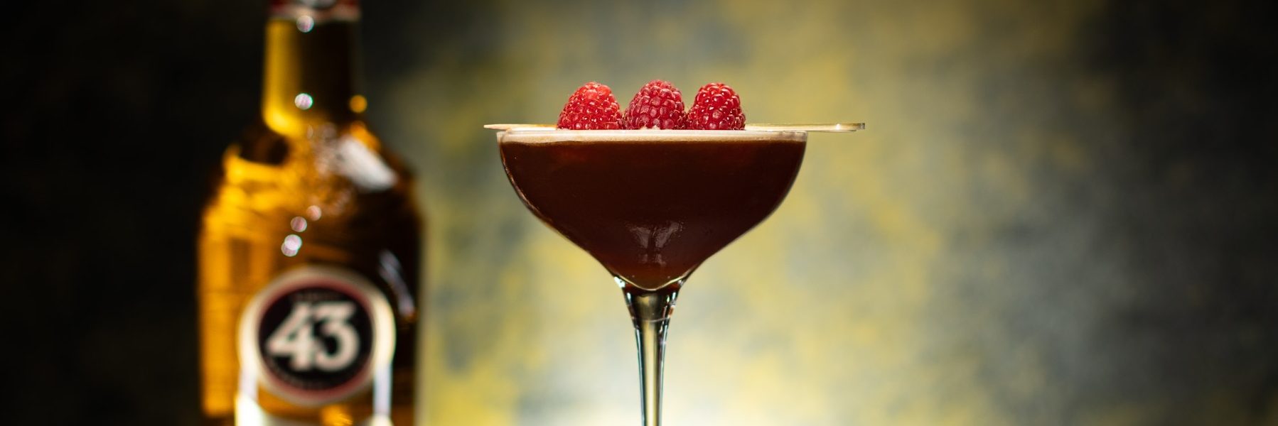 Licor 43 Deja Brew 43 in a coupe glass garnished with raspberries with licor 43 on yellow and black background